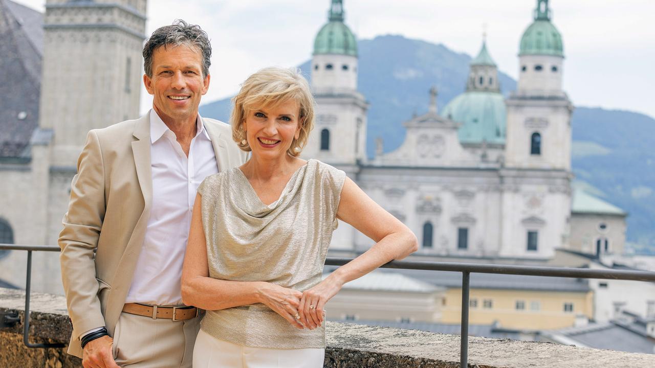Martin Ferdiny und Romy Seidl-Laux stehen vor der Kulisse historischer Kirchen und Häuser in der Altstadt von Salzburg.