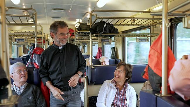 In einem älteren, gut gefüllten Zugabteil steht Josef Gründwidl mit dunklem Hemd und weißem Kollar zwischen sitzenden Fahrgästen, während Taschen und Jacken in den Gepäckablagen verstaut sind.