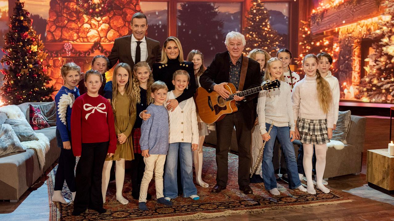 Kai Pflaume, Helene Fischer und Rolf Zuckowski mit Gitarre stehen mit Kindern vor einem funkelnden, gemütlich dekorierten Weihnachtskulissen‑Wohnzimmer.