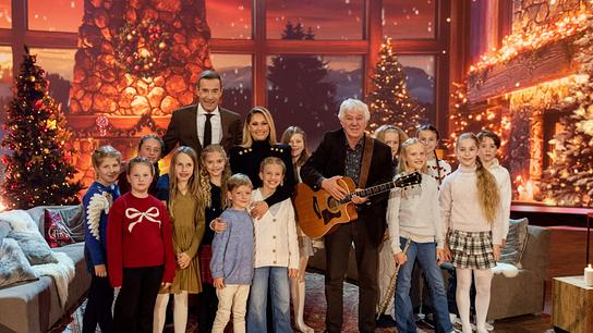 © ORF/NDR/Thorsten Jander Kai Pflaume, Helene Fischer und Rolf Zuckowski mit Gitarre stehen mit Kindern vor einem funkelnden, gemütlich dekorierten Weihnachtskulissen‑Wohnzimmer.