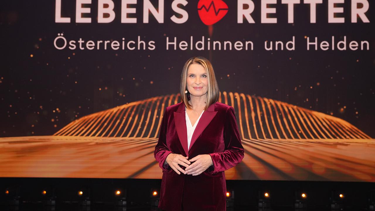 Barbara Stöckl im dunkelroten Samtanzug auf der Studiobühne mit Schriftzug "Lebensretter - Österreichs Heldinnen und Helden" im Hintergrund.