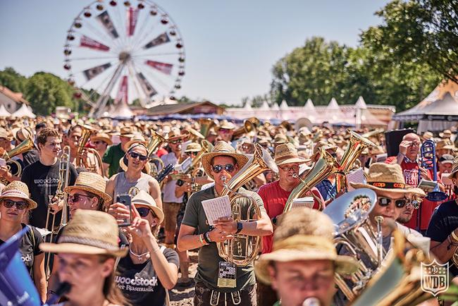 Impressionen vom „Woodstock der Blasmusik 2024“: Eine große Gruppe von Blasmusikerinnen und ‑musikern auf einem Festivalgelände, die im Freien spielen, mit einem Riesenrad im Hintergrund.