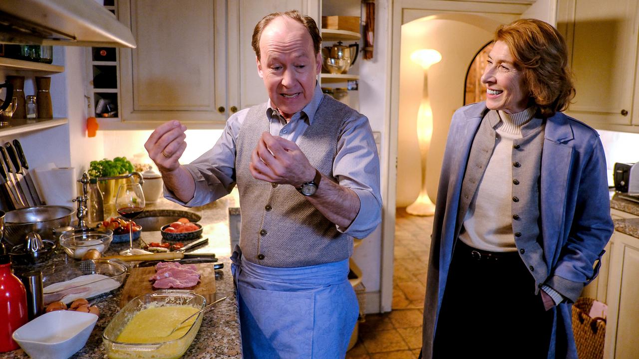 In einer Küche steht Anton beim Kochen an der Arbeitsplatte und gestikuliert, während Constanze daneben steht und lächelt.