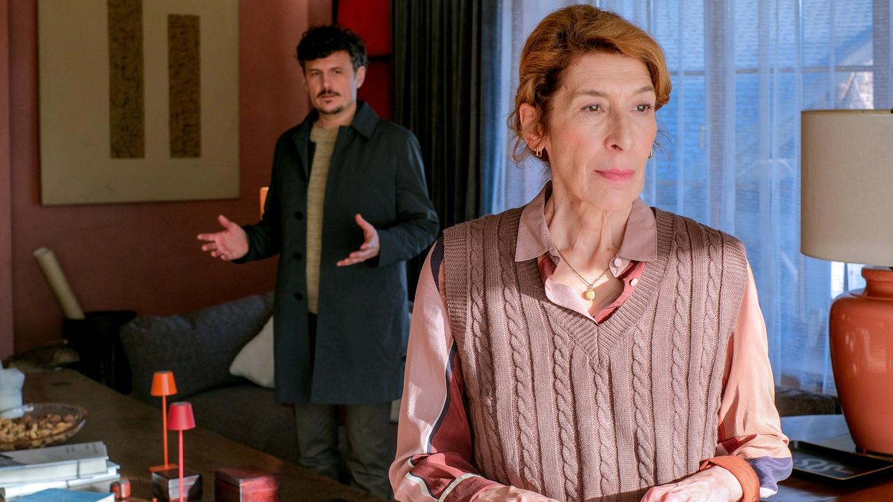 In einem Wohnzimmer steht Constanze mit einer gefalteten Zeitung in der Hand, während Ricardo hinter ihr stehend im Hintergrund gestikulierend spricht.