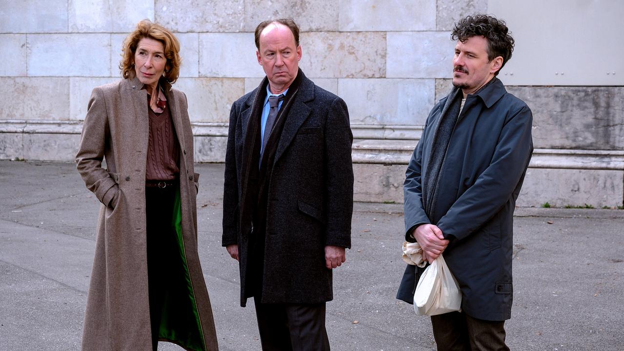 Constanze, Anton und Ricardo  stehen ernst blickend im Freien nebeneinander vor einer Steinmauer.
