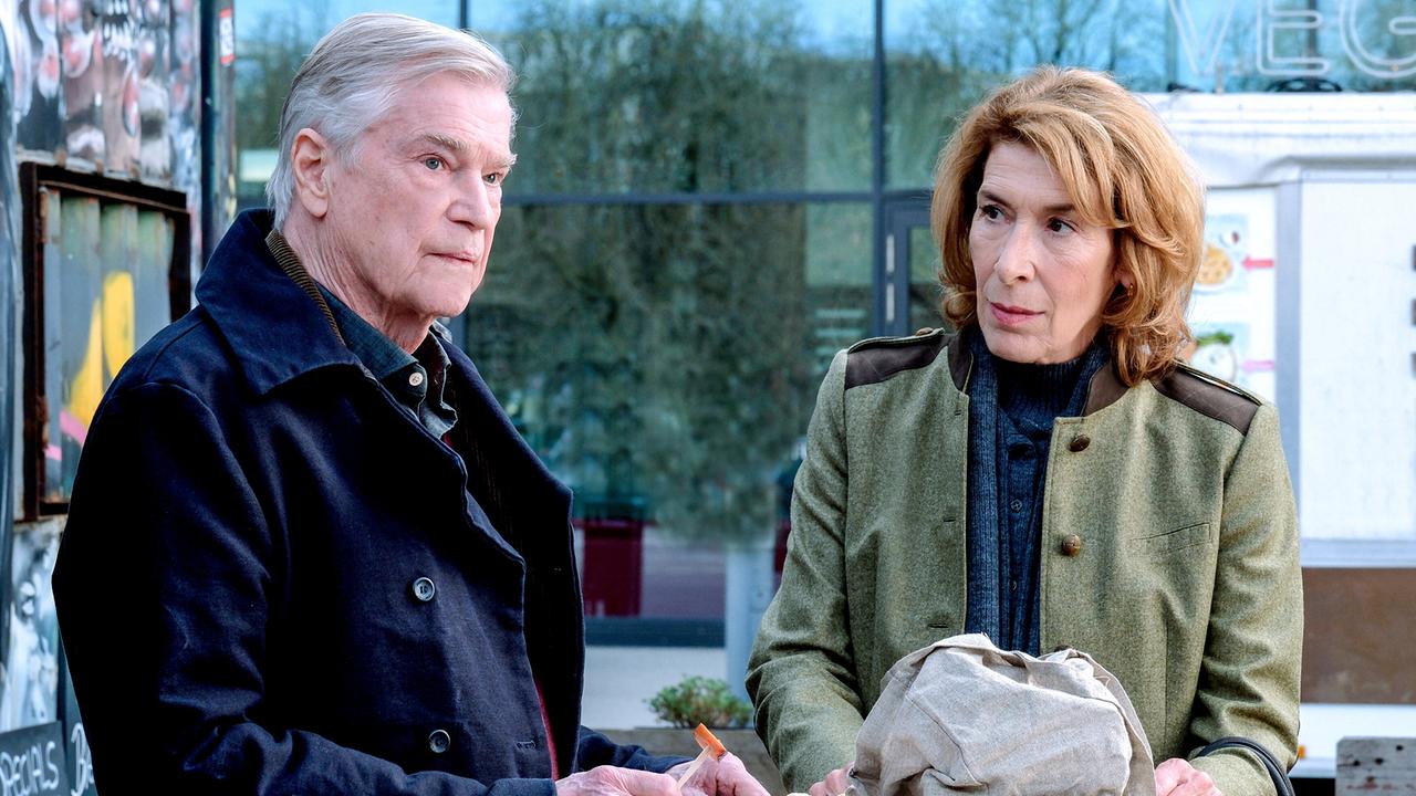 An einem kleinen Stehtisch im Freien schreibt der ältere Richard Westerhoff etwas auf, während Constanze daneben ihn anblickt.