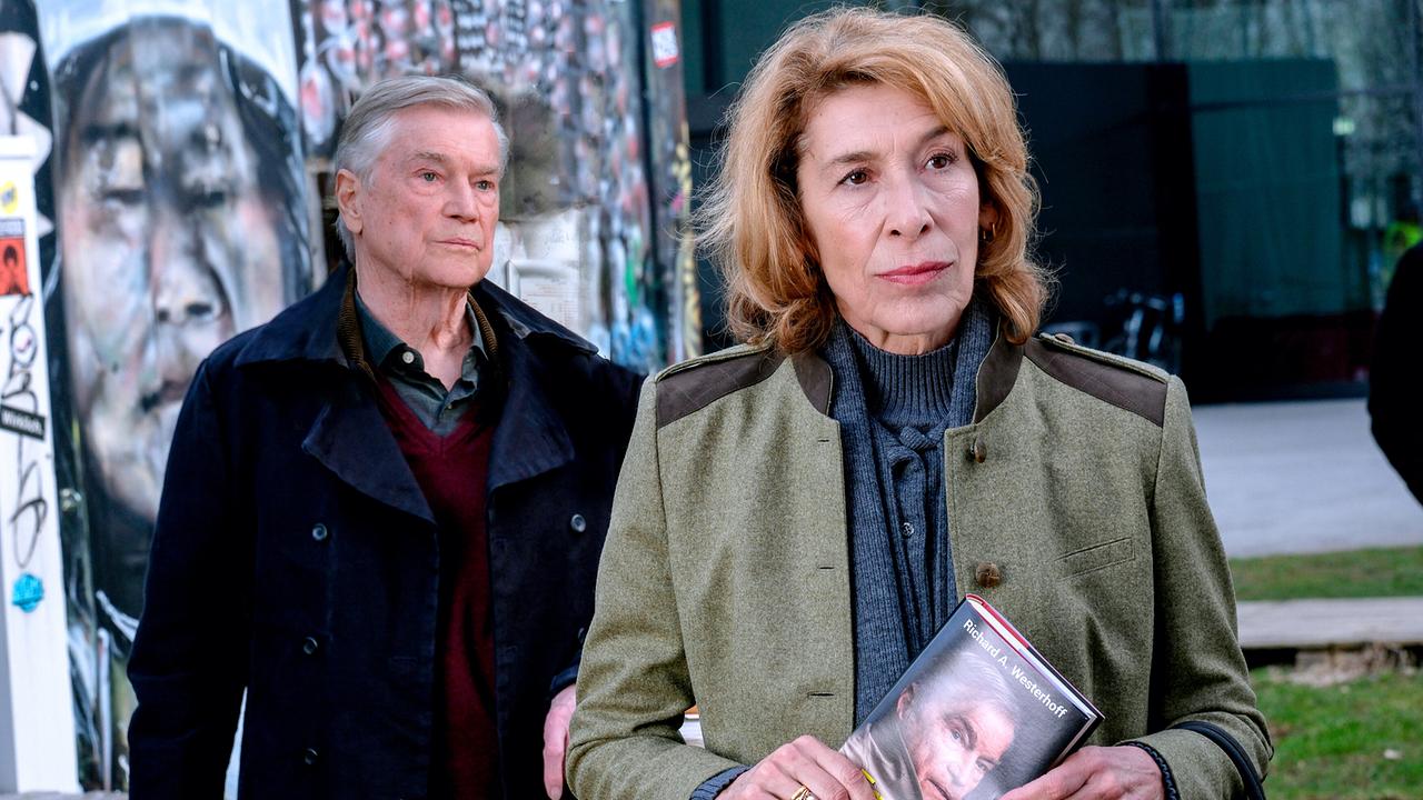 Vor einer Graffiti-Wand im Freien steht Constanze Laux mit einem Buch von Richard Westerhoff in der Hand, während Richard Westerhoff hinter ihr steht.