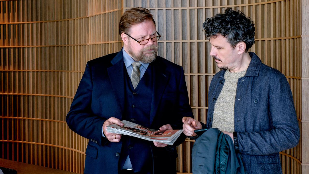 In einem Foyer zeigt Portier Josef Ricardo Westerhoff ein Magazin vor einer Holzpanelwand.