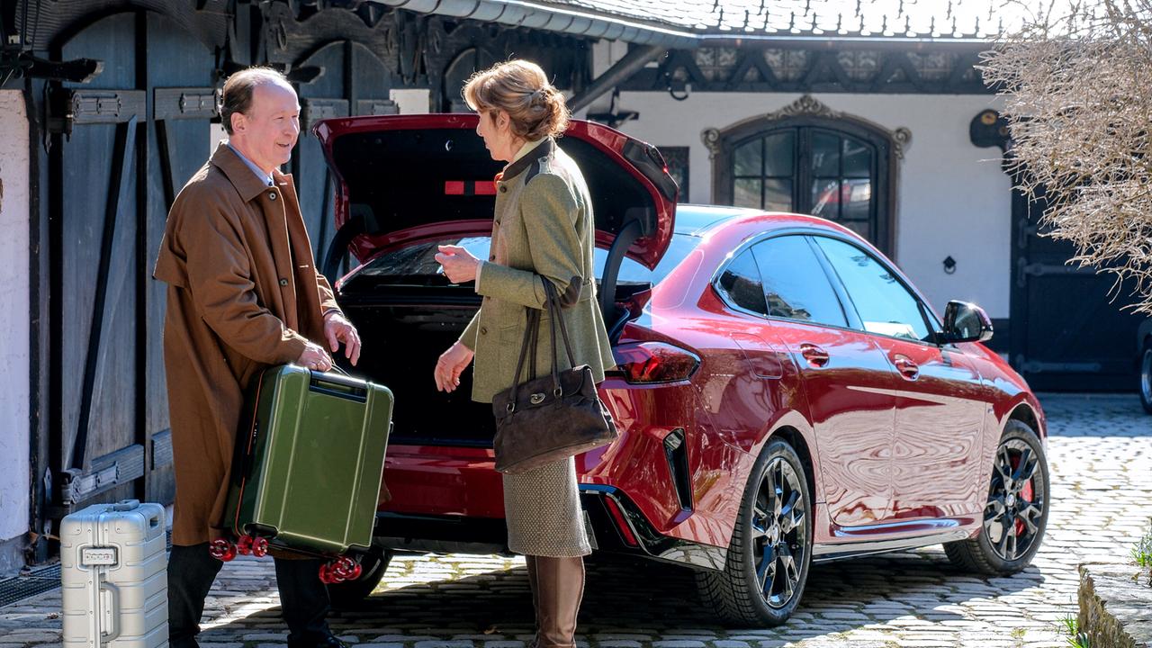 Vor einem Haus steht Anton Laux, mit einem Koffer in der Hand, neben Constanze Laux, dahinter ein dunkelroter Wagen mit geöffnetem Kofferraum.