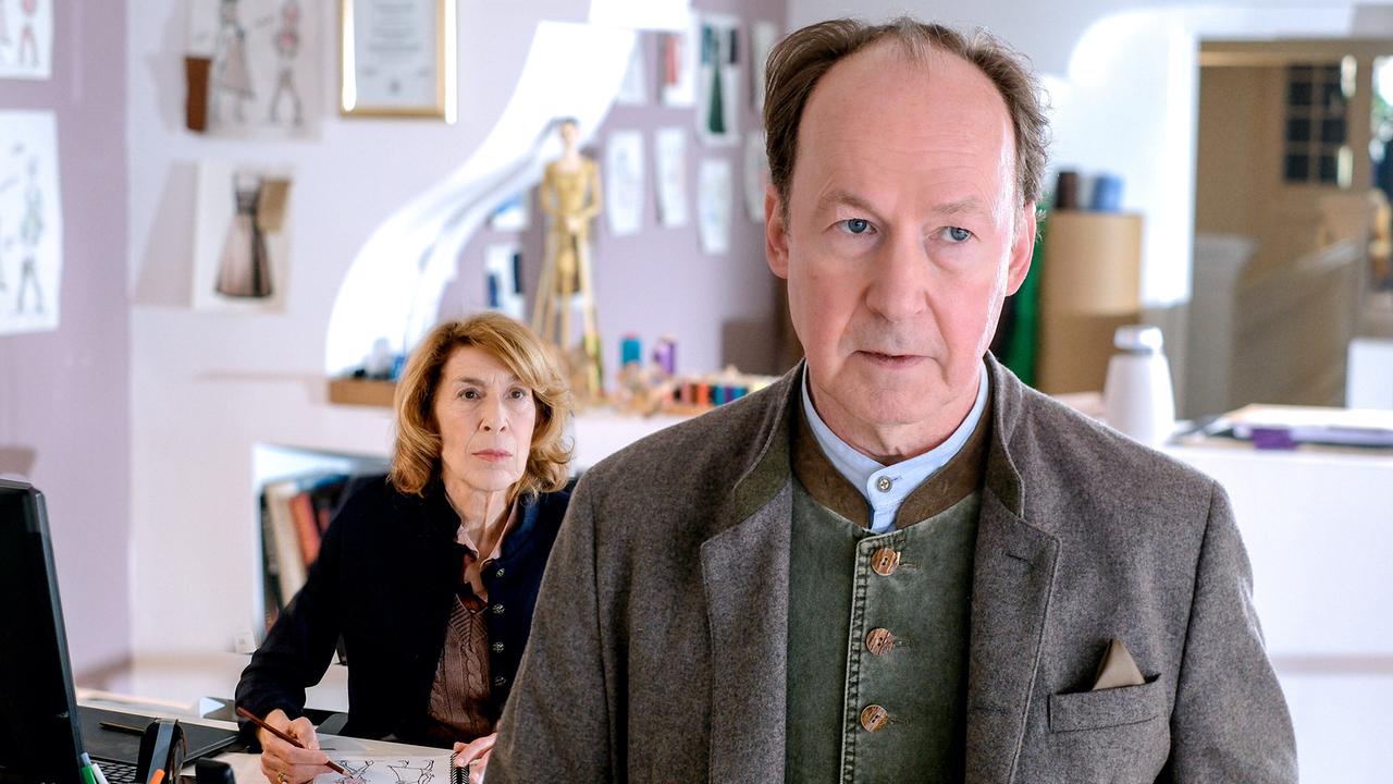 Anton Laux steht in einem hellen Wohnraum und blickt nach vorne, während im Hintergrund Constanze Laux am Schreibtisch an Entwürfen arbeitet und zu ihm schaut.