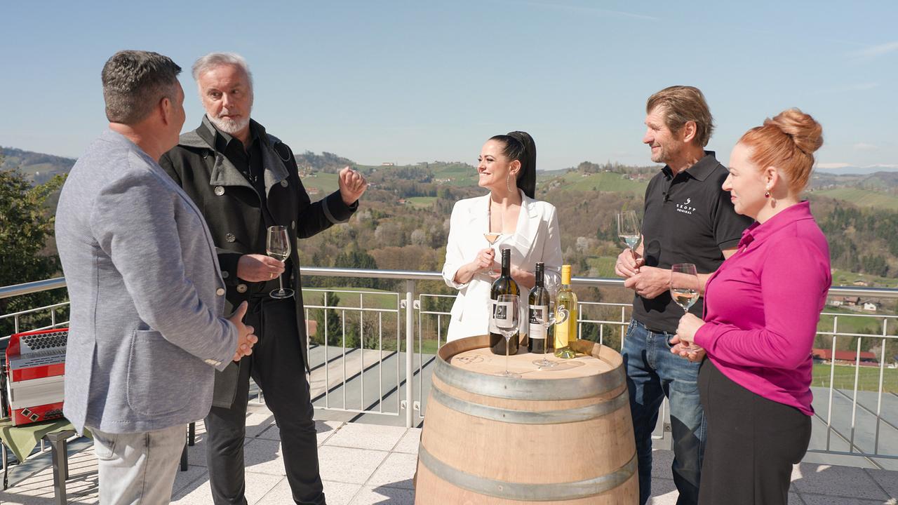© ORF/televisionair Marc Pircher, Fred Jaklitsch, Jeannine Rossi, Walter Skoff und Eva Skoff-Liebau stehen auf einer Terrasse um ein Weinfass und verkosten Wein mit Blick ins Hügelland.