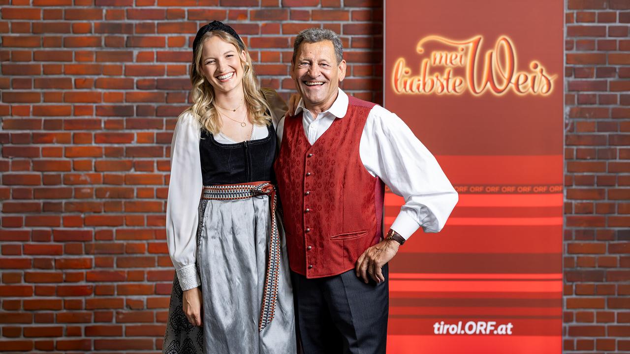 © ORF/ORF-Tirol/Thomas Steinlechner Diana Foidl und Franz Posch posieren lachend vor einer Backsteinwand und einem ORF‑Roll‑Up mit dem Schriftzug „Mei liabste Weis“.