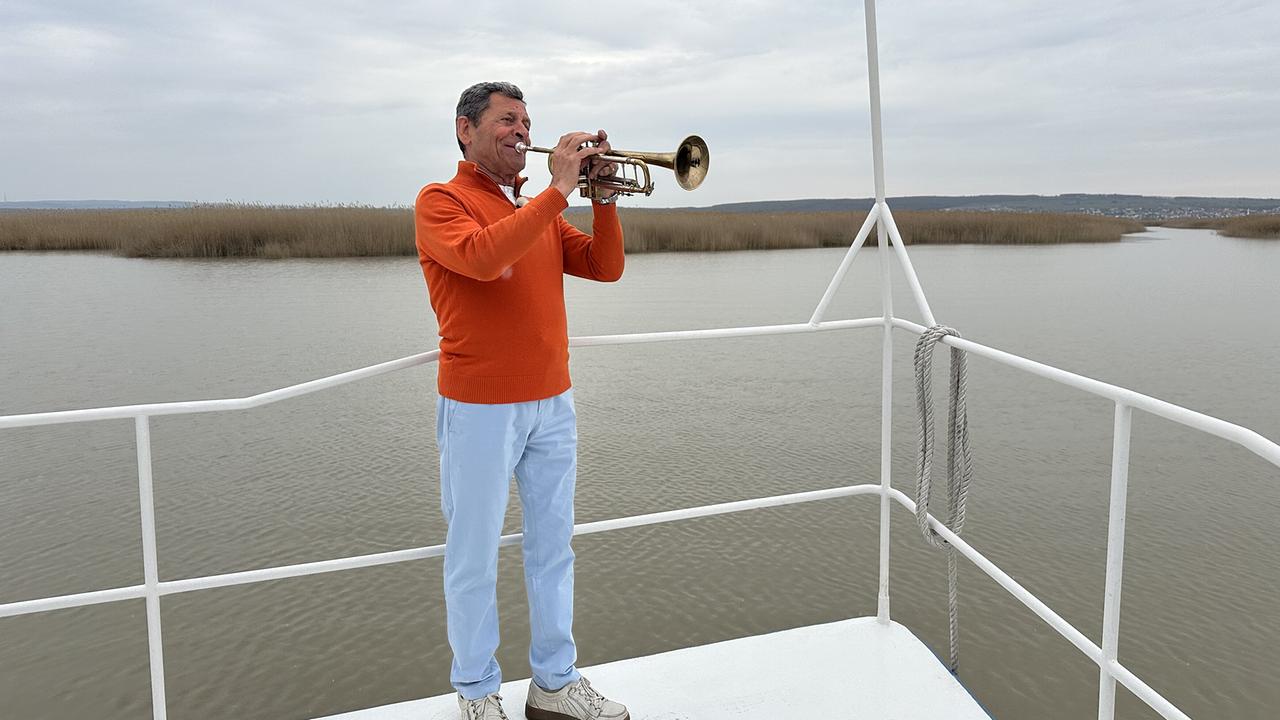 Franz Posch gibt das Signalhorn auf dem Deck eines Boots vor einer ruhigen Seenlandschaft.