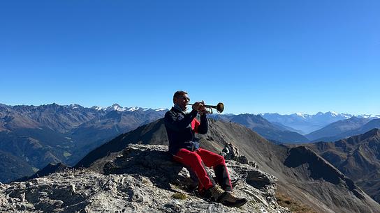 Franz Posch spielt Trompete auf einem felsigen Gipfel in der beeindruckenden Berglandschaft von Serfaus-Fiss-Ladis.