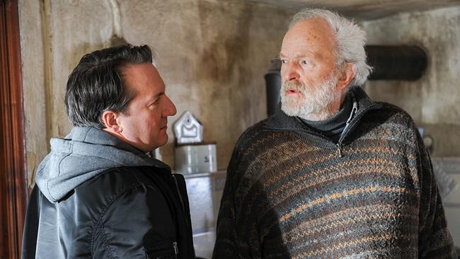 Zwei Personen stehen in einer alten Küche unterhalten sich, im Hintergrund sind schmutzige Wände und ein Ofenrohr zu sehen