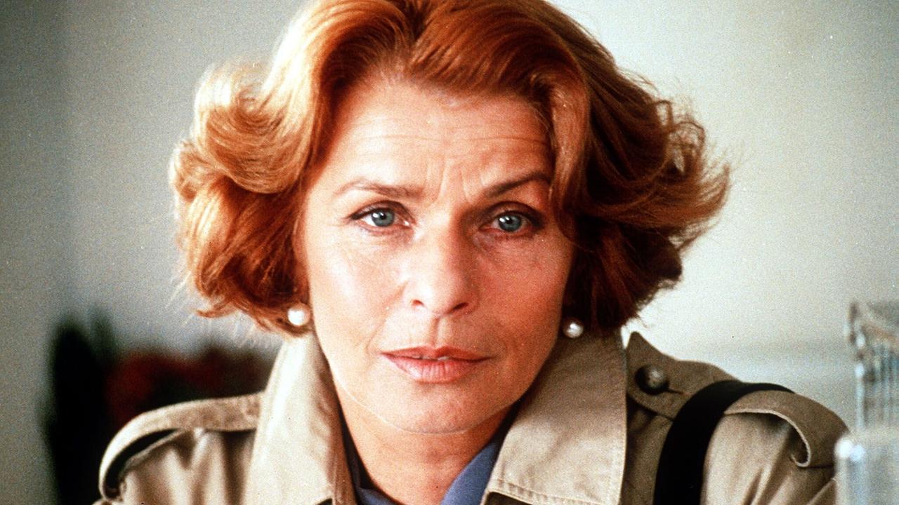 Nahaufnahme von Senta Berger mit rötlichem Haar und hellem Mantel in einem neutral beleuchteten Innenraum.