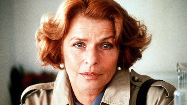 Nahaufnahme von Senta Berger mit rötlichem Haar und hellem Mantel in einem neutral beleuchteten Innenraum.