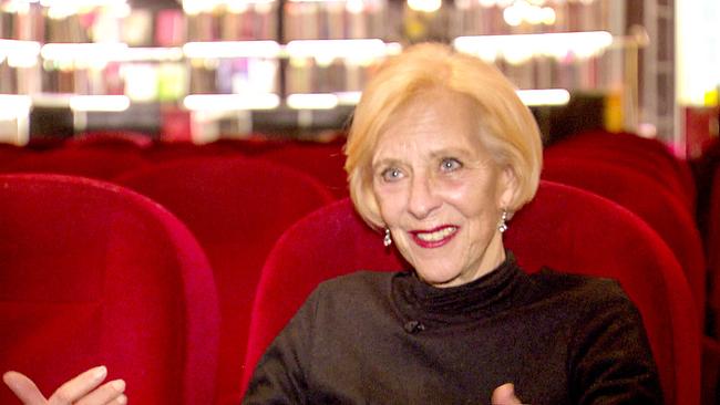 Käthe Kratz sitzt in einem Kinosaal zwischen roten Polstersitzen und spricht gestikulierend, während im Hintergrund die leeren Reihen des Zuschauerraums zu sehen sind.