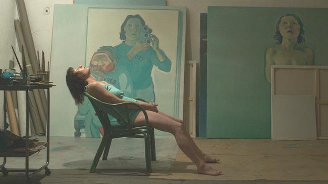 © ORF/coop99 filmproduktion Birgit Minichmayr als Maria Lassnig lehnt fast liegend in einem Atelier in einem Stuhl, während im Hintergrund mehrere große Gemälde und aufgestellte Leinwände an der Wand stehen.