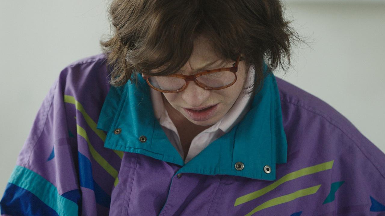 Nahaufnahme von Birgit Minichmayr als Maria Lassnig in einer lila-türkisen Jacke, die nach unten blickt.