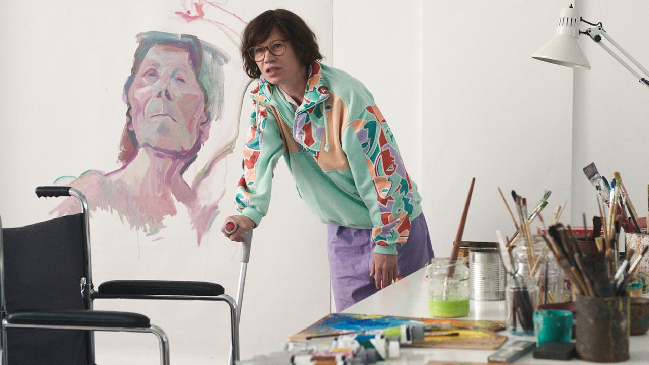 In einem Atelier steht Maria Lassnigmit Krücke neben einem Tisch voller Pinsel und Malutensilien, im Hintergrund ein großes Frauenporträt.