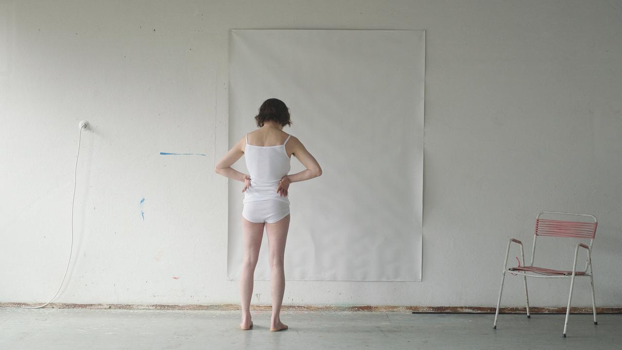 Maria Lassnig steht in weißer Unterwäsche vor einer großen, leeren Leinwandfläche an der Wand, daneben steht ein Metallsessel.