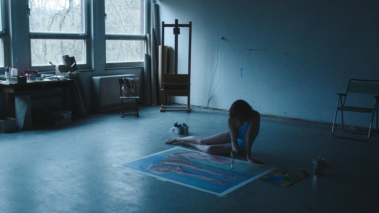 In einem dunklen Atelier sitzt Maria Lassnig am Boden und malt an einem großformatigen Bild, während Tageslicht durch große Fenster fällt.