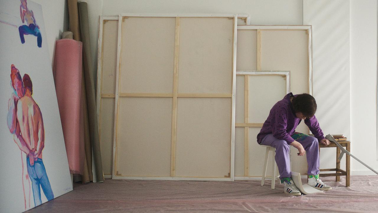 In einem Atelier sitzt Maria Lassnig in bunter Kleidung auf einem Hocker, neben ihr lehnt eine Armkrücke auf einem kleinen Tisch, an der linken Wand lehnt ein Gemälde, hinter stehen große, bespannte Leinwände.