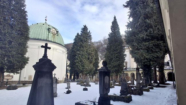 © ORF/Landesstudio Salzburg Ein verschneiter historischer Friedhof mit dunklen Grabmälern liegt zwischen hohen Immergrünen und einer Kirche mit grüner Kuppel.
