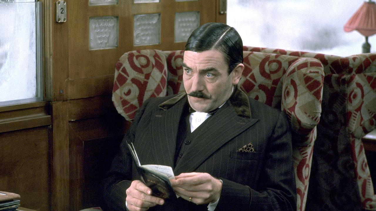 © ORF/Studiocanal Meisterdetektiv Hercule Poirot mit streng gescheitelter Frisur und markantem Schnauzbart, trägt einen Anzug und sitzt im Zug in einem Lehnsessel und hat sein Notizbuch in den Händen