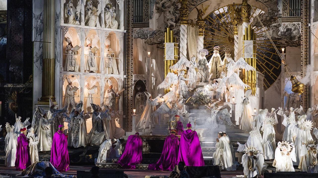 Opernszene aus der "Tosca" aus dem Steinbruch St. Margarethen mit zahlreichen Darstellerinnen und Darstellern in weißen, religiös anmutenden Kostümen und mehrere Darsteller:innen in leuchtend violetten Mänteln.