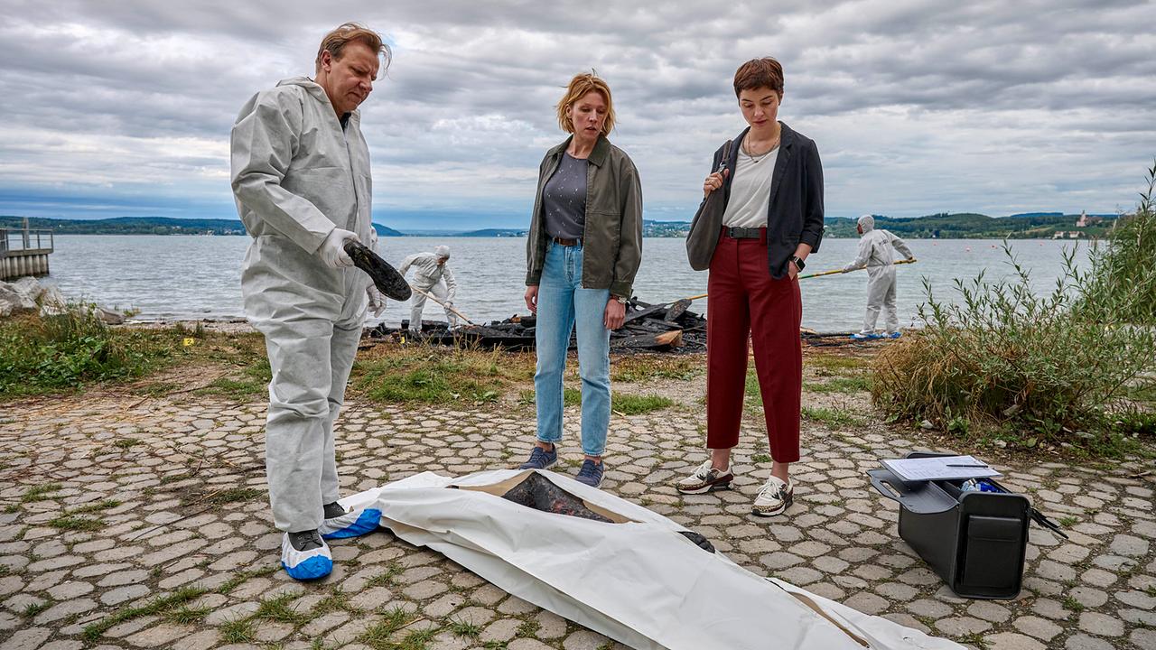© ORF/ZDF/Patrick Pfeiffer Konstanz Gerichtsmediziner Gregor Eisen im Schutzanzug gibt den Kriminalkommissarinnen Doro Beitinger und Chiara Locatelli am Tatort am Ufer des Bodensees neben einem abgedeckten Leichnam erste Informationen zum brisanten Todesfall.