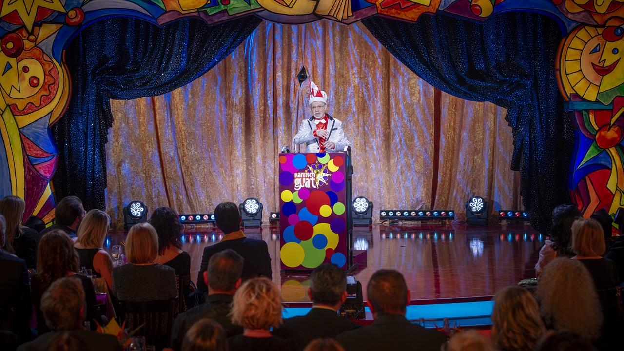 Alexius Vogel in festlichem Kostüm steht hinter einem bunt gestalteten Rednerpult auf der bunt gerahmten "Narrisch guat"-Bühne, während das Publikum davor sitzt.