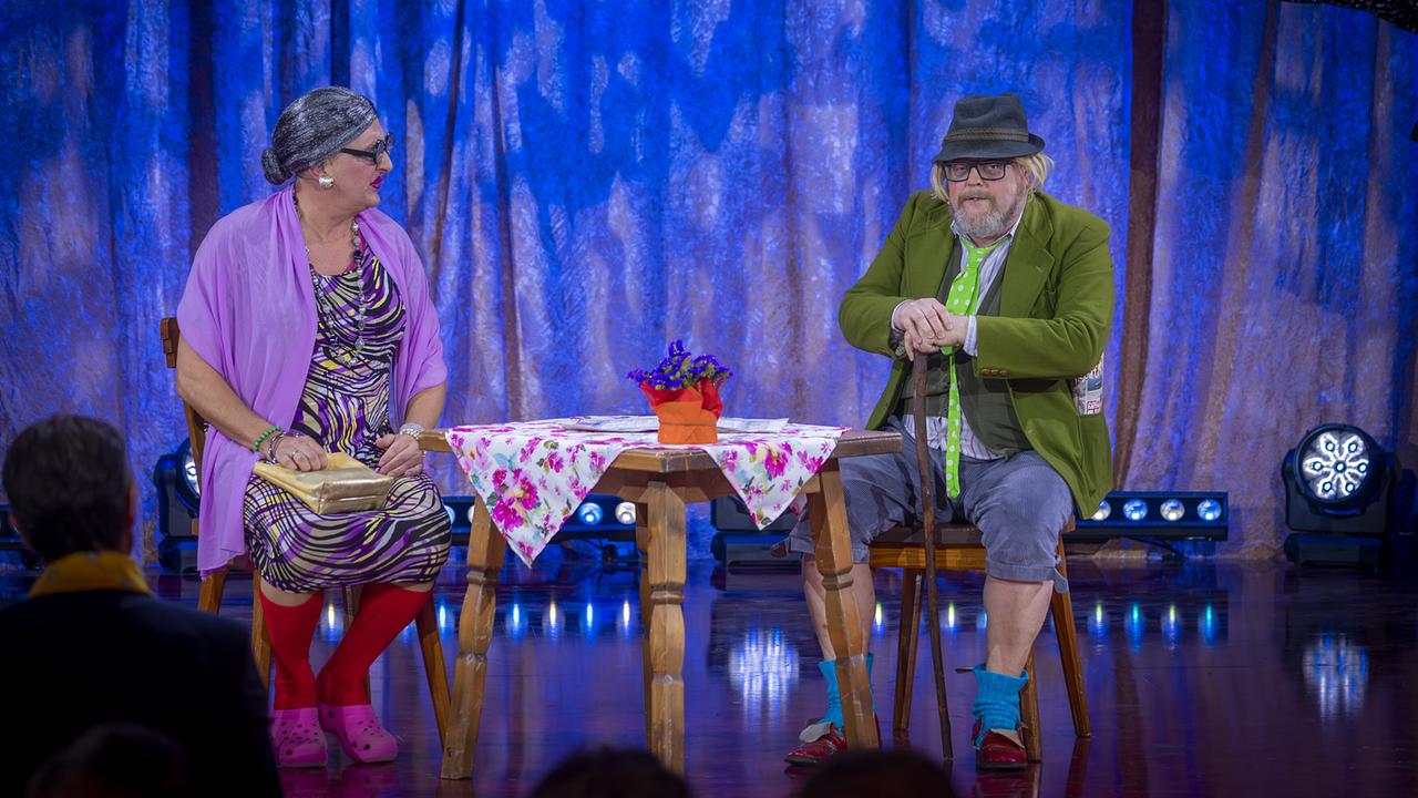 „Der Tschentsche und seine Henriette“ dargestellt von Andreas Fischer und Willie Wurzer sitzen an einem kleinen Tisch mit Blumen auf der "Narrisch guat"-Bühne vor Publikum. 