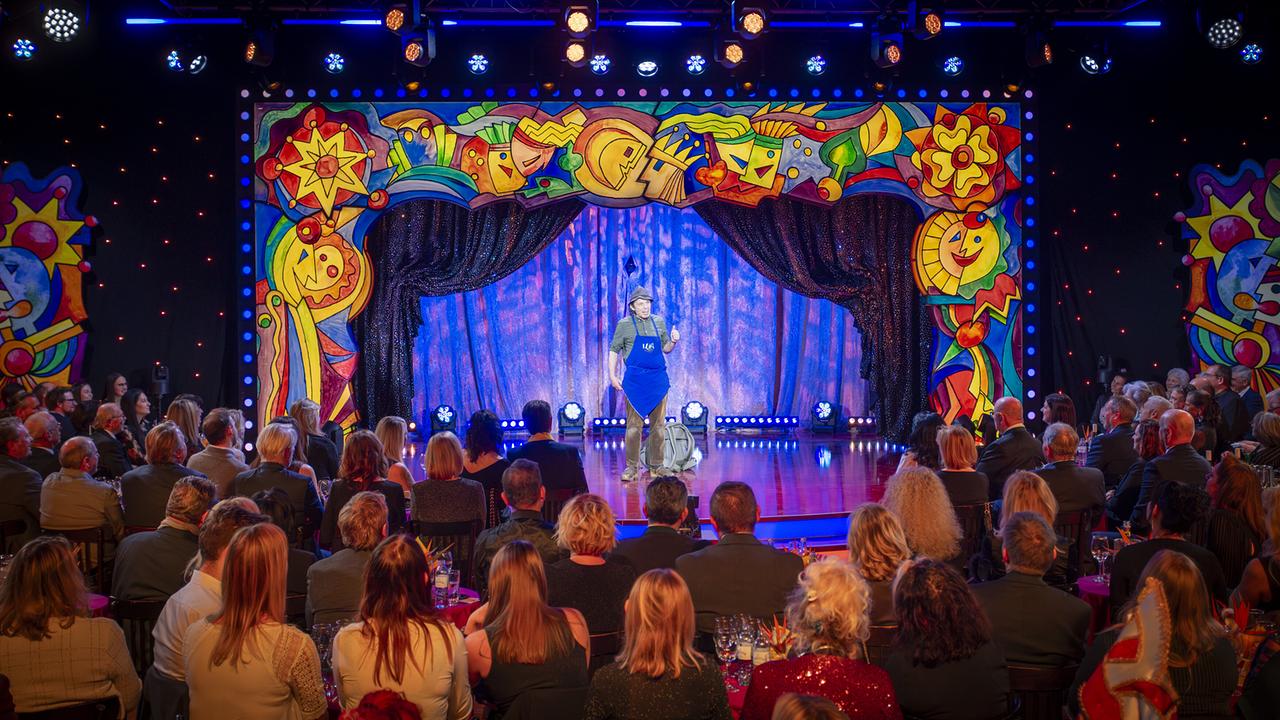 Luis aus Südtirol mit Hut, kariertem Hemd und einer blaue Schürze mit der Aufschrift „Luis Südtirol“ steht auf der bunt gerahmten "Narrisch guat"-Bühne vor Publikum.