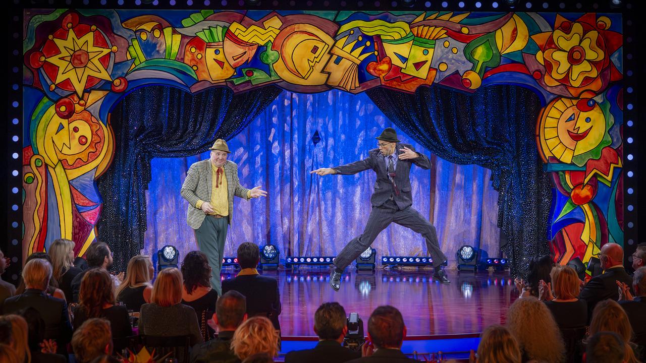 Christopher Visotschnig und Reno Paulic stehen auf der bunt gerahmten "Narrisch guat"-Bühne bei ihrem Stück "Nach der Messe", während Reno Paulic in Anzug und Hut dynamisch in die Luft springt.