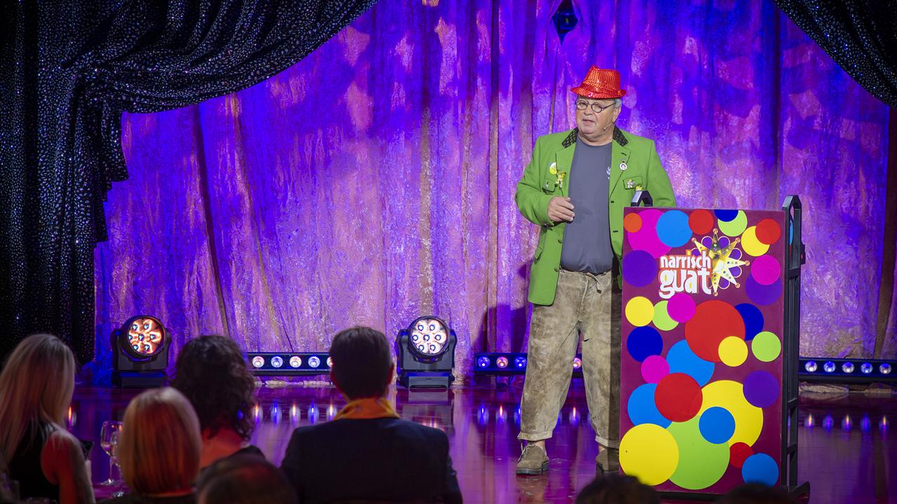 Sepp Wölbitsch in grünem Sakko und rotem Hut steht neben einem bunten Rednerpult vor Publikum auf der "Narrisch guat"-Bühne.