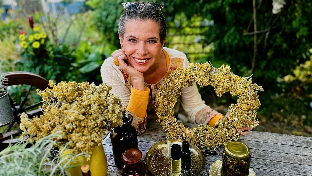 Uschi Zezelitsch hält einen Immortelle-Kranz am Holztisch, umgeben von Kräutern und Ölen in Glasgefäßen im Freien.