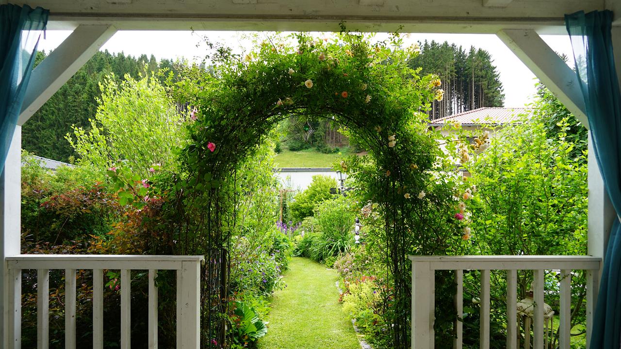 © Leopold Mayrhofer/Natur im Garten/Leopold Mayrhofer Romantischer Gartendurchgang mit Rosenbogen, Rasenweg und bunten Staudenbeeten, gerahmt von weißem Pavillon