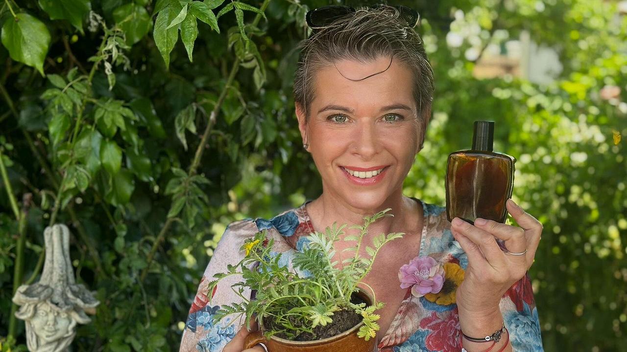 Uschi Zezelitsch steht im Garten und hält einen braunen Topf mit Gänsefingerpflanze und eine braune Flasche Rasierwasser.
