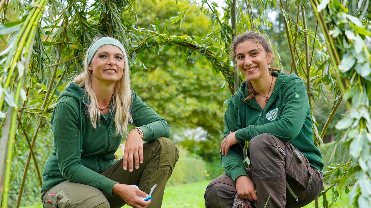 © Leopold Mayrhofer/Natur im Garten Zwei lächelnde Gärtnerinnen in grünen Jacken knien in einem fertigen Tipi aus grünen Weidenruten.