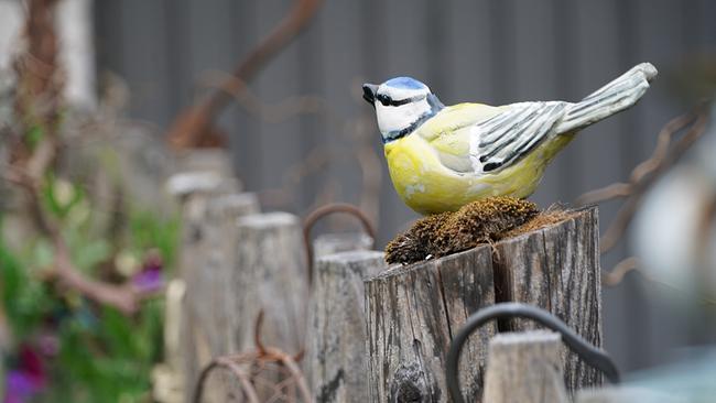 Keramikfigur einer Blaumeise mit gelber Brust sitzt auf einem verwitterten Holzpfosten im Garten.