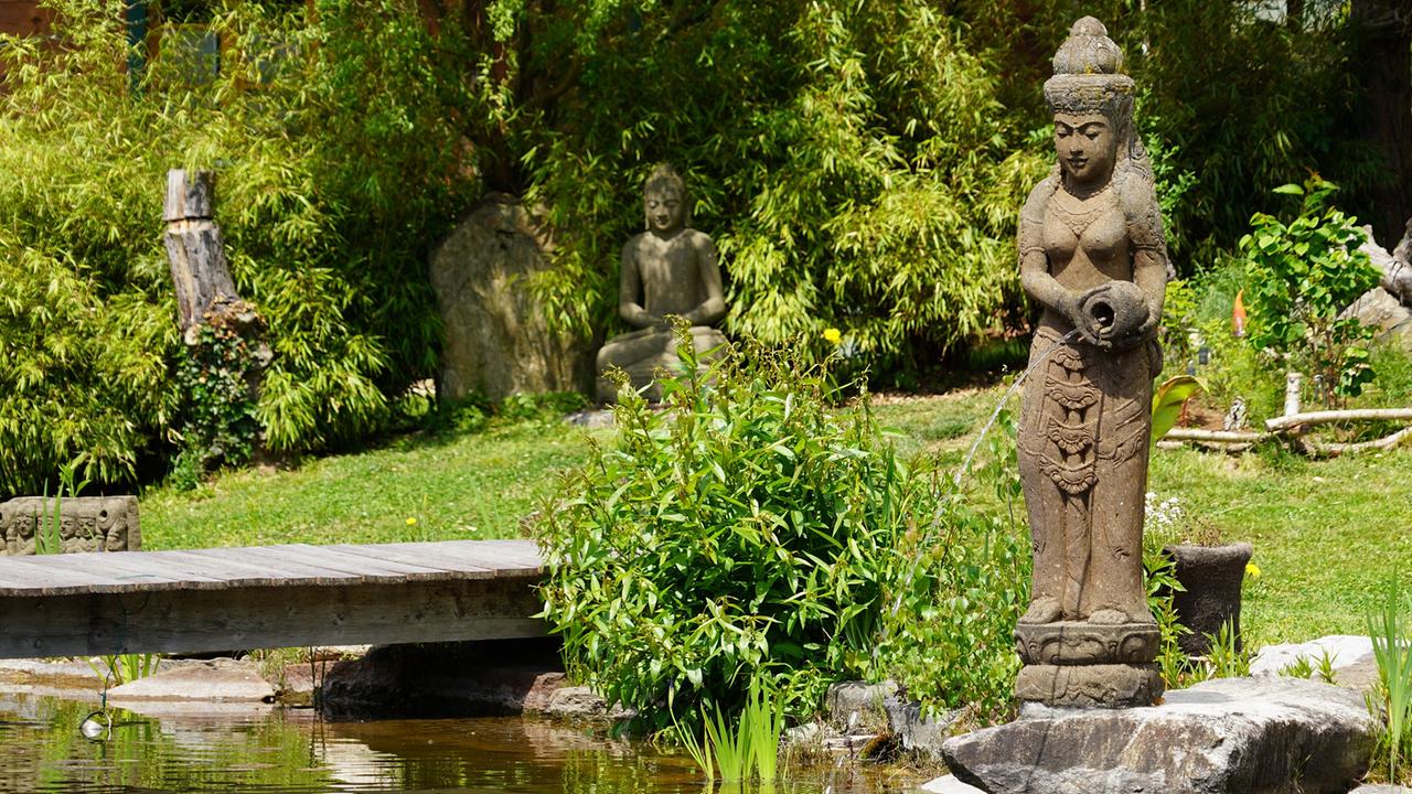 © Leopold Mayrhofer/Natur im Garten Steinfigur mit asiatischen Merkmalen steht am Rand eines Teichs in einem fernöstlich gestalteten Naturgarten.