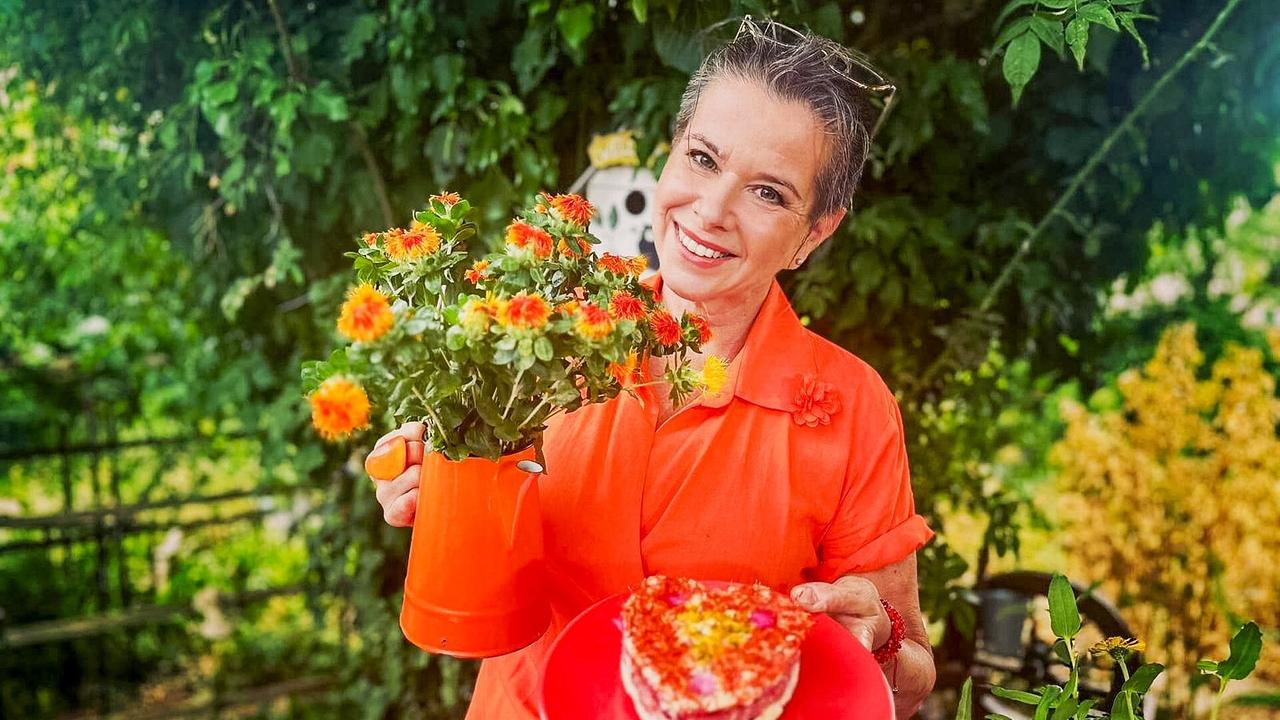 © ORF/Uschi Zezelitsch Uschi Zezelitsch steht im Garten und hält eine orange Gießkanne mit Färberdisteln und einen roten Teller mit Kuchen.