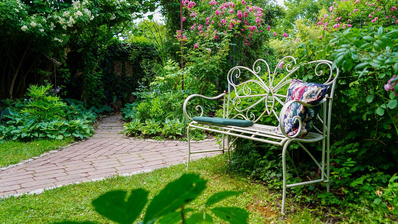 © Leopold Mayrhofer/Natur im Garten Eine Gartenbank mit floral gemustertem Kissen im blühenden, grünen Garten von Martina und Haimo Ungersböck.