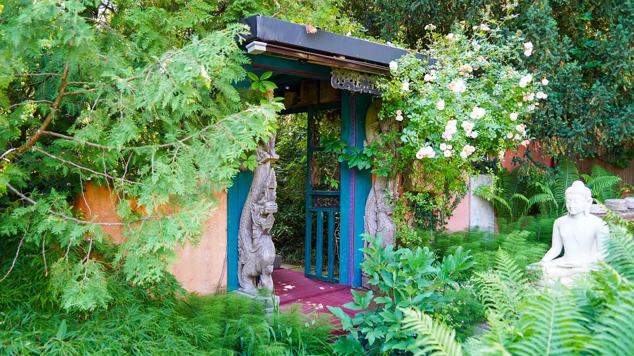 © Leopold Mayrhofer/Natur im Garten Gartentor in asiatischem Stil mit geschnitzten Holzfiguren, umgeben von Farnen, Rosen und üppigem Grün.