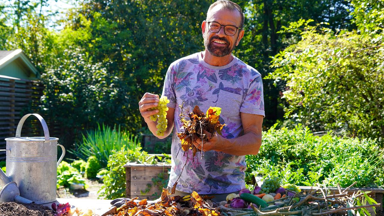 © Leopold Mayrhofer/Natur im Garten Karl Ploberger steht an einem Gartentisch und zeigt Weintrauben, Laub und Pflanzenreste für Kompost im sonnigen Garten