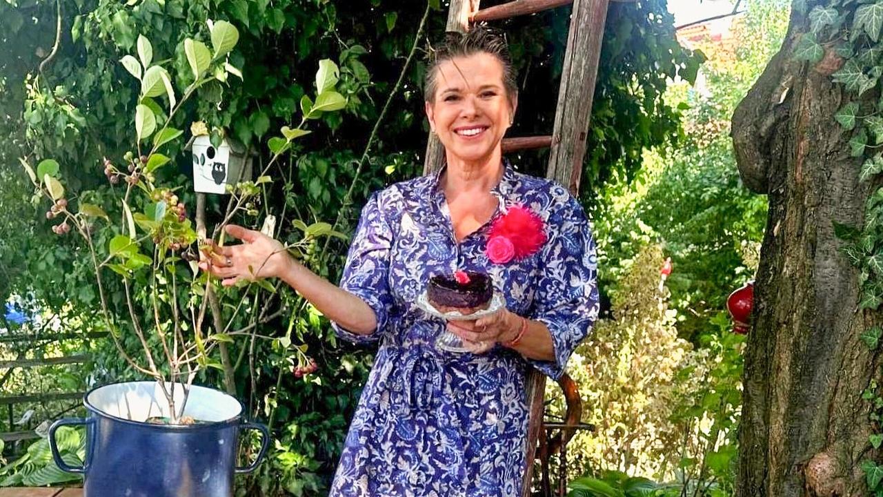 © ORF/Uschi Zezelitsch Uschi Zezelitsch hält ein Glas mit Superwoman-Kuchen aus Aroniabeeren und steht neben eineR Aroniapflanze im Garten: