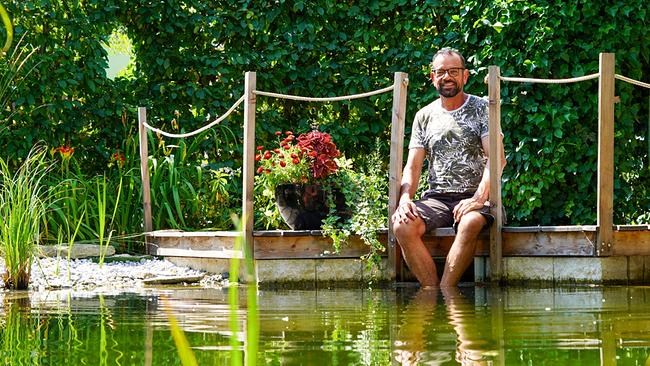 © Leopold Mayrhofer/Natur im Garten Karl Ploberger sitzt entspannt mit den Füßen im Wasser auf einem Holzsteg am Teich.