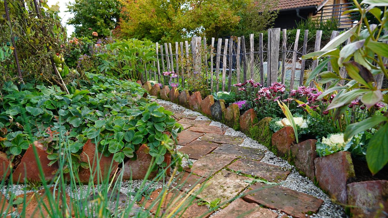 Gartenweg aus Steinplatten auf Kies, links davon grüne Pflanzen, rechts ein niedriger Zaun mit bunten Blumen